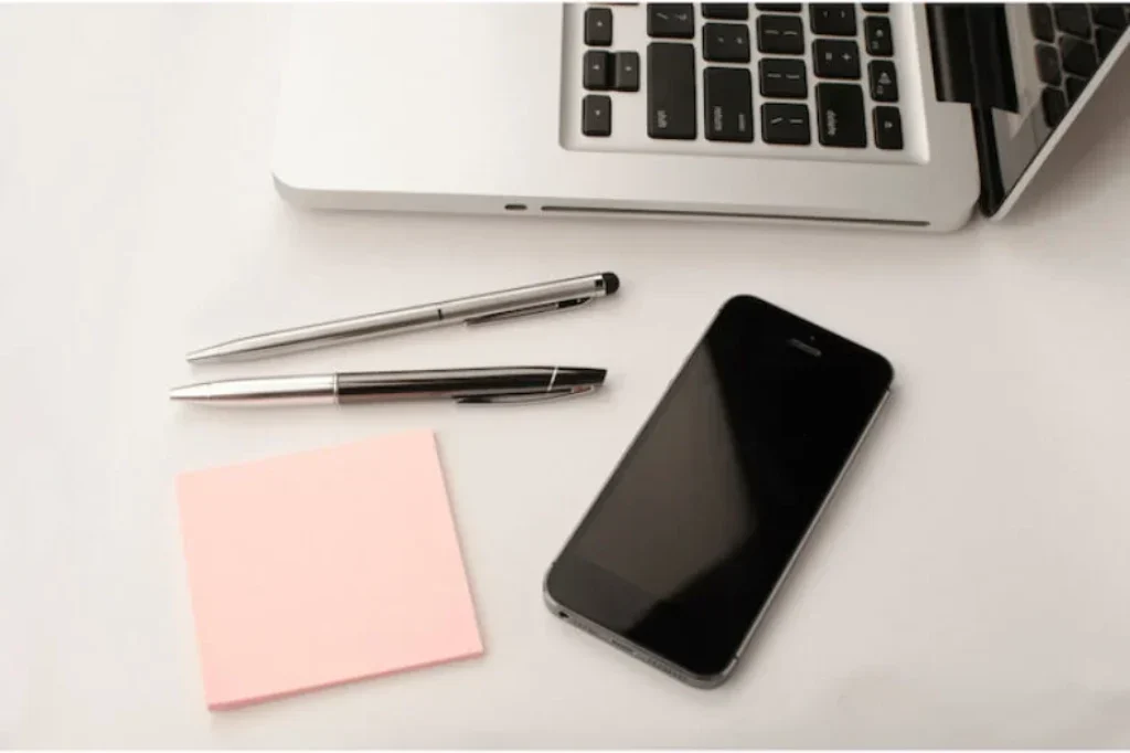 Uma mesa que contém um notebook, um smartphone, um papel e duas canetas.