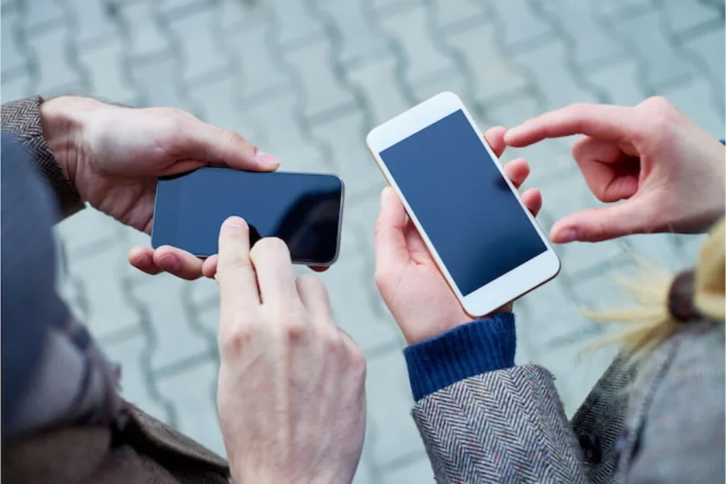 Duas pessoas segurando smartphones em mãos, interagindo com as telas touch, com fundo de calçamento urbano desfocado.
